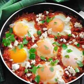 A skillet of shakshuka with poached eggs in tomato sauce, topped with crumbled cheese and fresh herbs, placed on a green cloth next to a dark wooden surface.