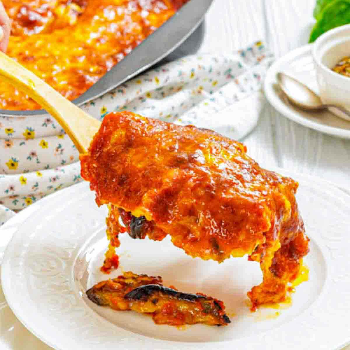 A slice of baked eggplant parmesan is being served onto a white plate, with a casserole dish and utensils in the background.