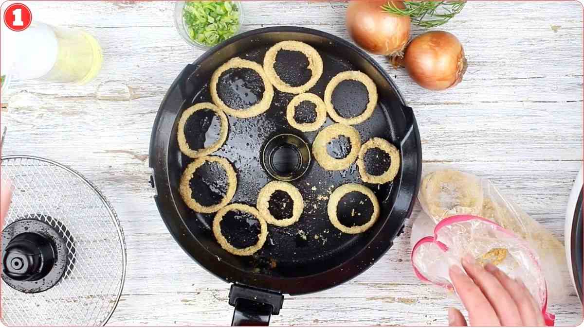 Slices of onion coated in batter are arranged in a single layer inside an air fryer basket, with onions and chopped green onions nearby on a light wooden surface.