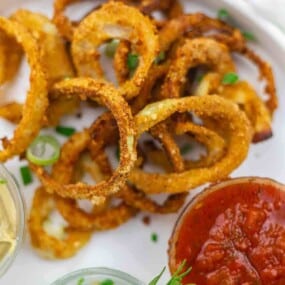 A plate of crispy fried onion rings garnished with green herbs, served with a side of red dipping sauce.
