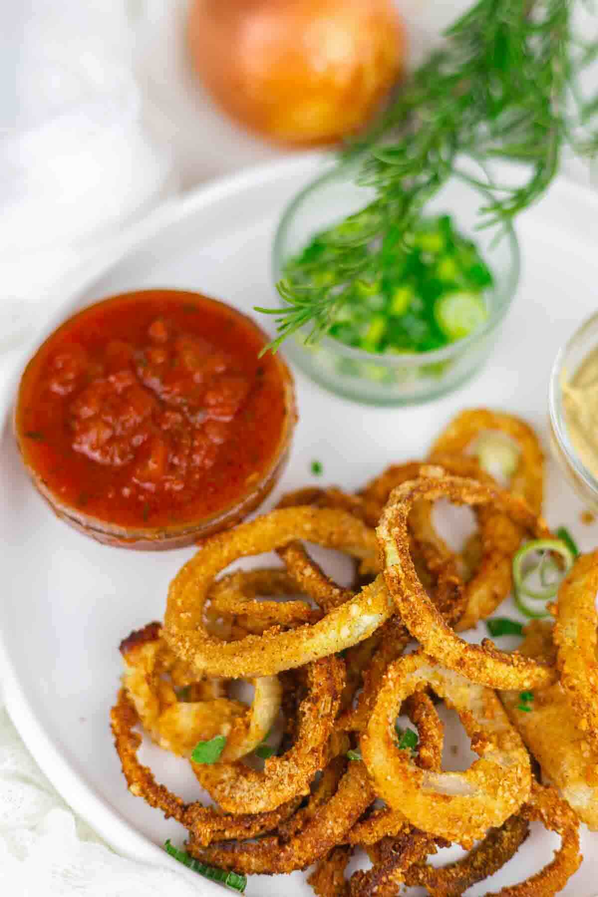 A plate of crispy fried onion rings served with salsa, chopped green onions, and a dip, garnished with a sprig of rosemary.