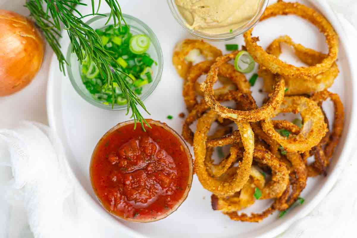 A plate of crispy onion rings with bowls of tomato salsa, green onions, fresh dill, and a creamy dipping sauce.