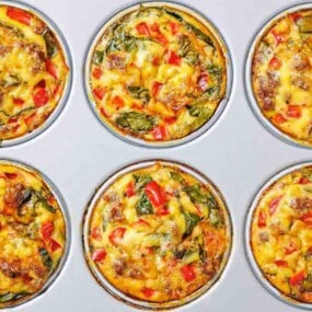 Six baked egg muffins with visible vegetables and sausage pieces in a metal muffin tin.