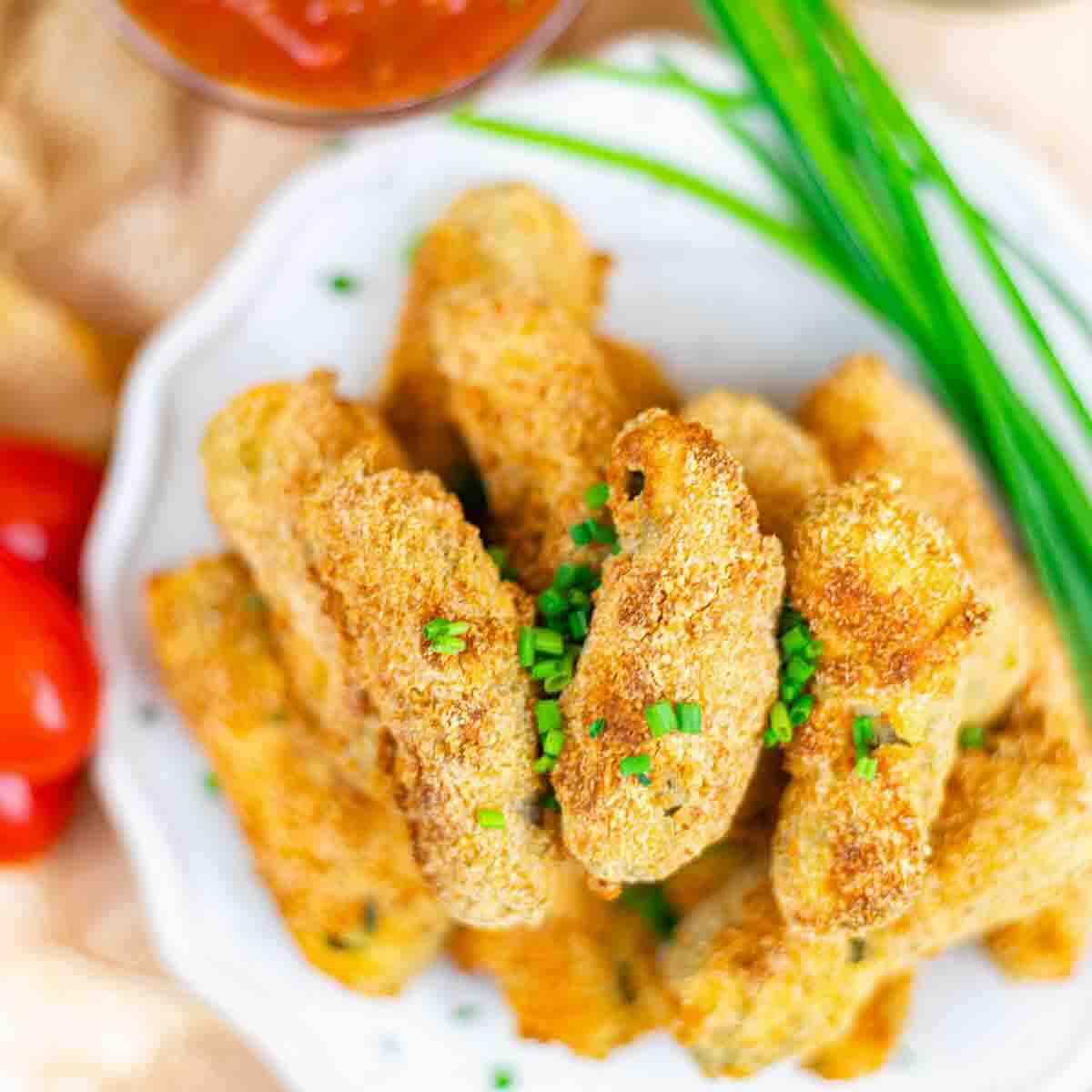 A plate of breaded, baked zucchini fries garnished with chopped chives, with a bowl of red dipping sauce and fresh green herbs in the background.