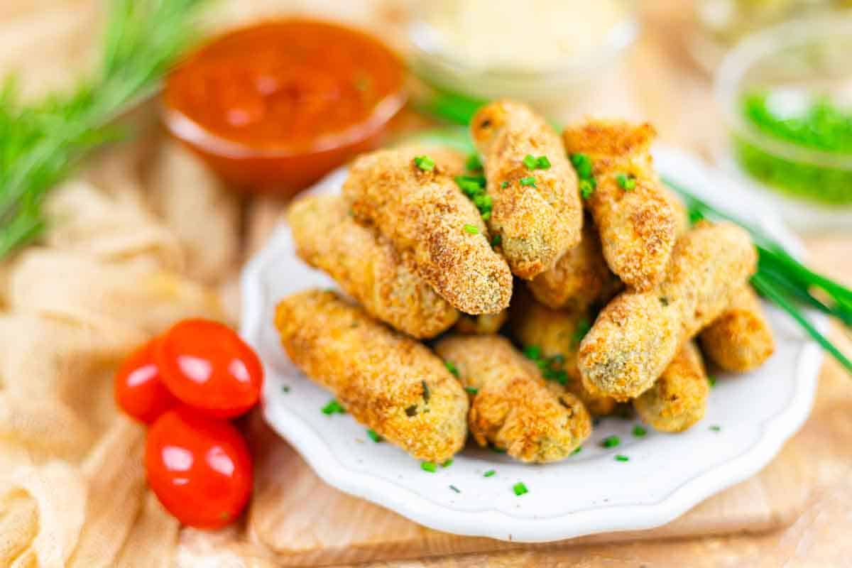 A plate of breaded, fried pickle sticks garnished with chopped chives, with bowls of dipping sauces and cherry tomatoes in the background.