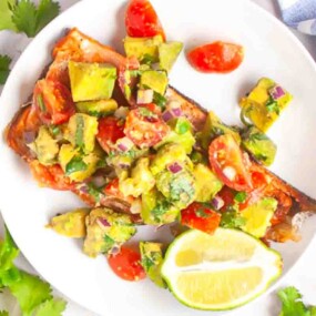 Grilled salmon fillet topped with avocado and tomato salsa, served with a lime wedge on a white plate.