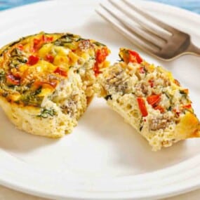 A mini frittata with vegetables and sausage, sliced in half on a white plate with a fork beside it.