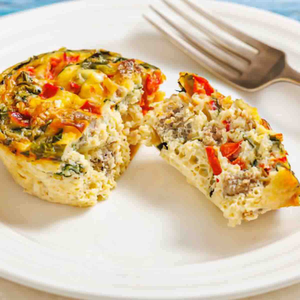 A mini frittata with vegetables and sausage, sliced in half on a white plate with a fork beside it.