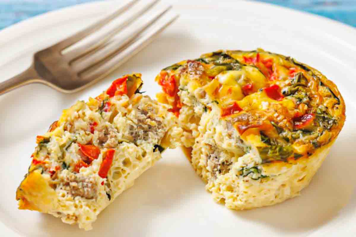 An egg muffin with visible sausage, red peppers, and greens is cut in half on a white plate beside a fork.