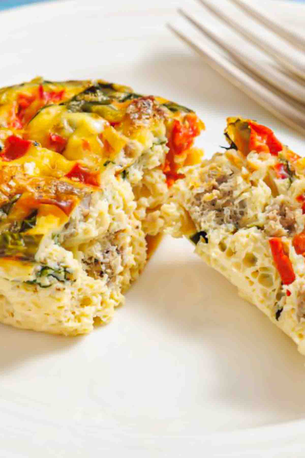 A slice of egg casserole with visible sausage, red bell peppers, and spinach on a white plate, next to a fork.