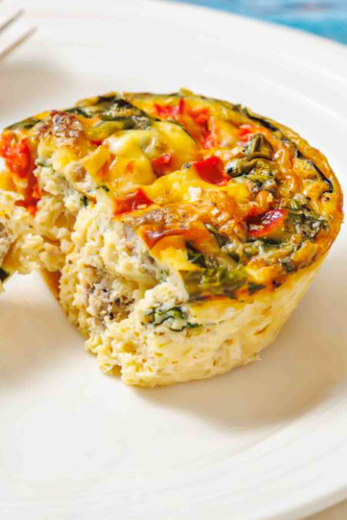 A baked egg muffin with vegetables and herbs sits on a white plate, with a bite taken out, revealing the inside texture.