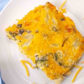 A slice of baked casserole with a golden cheese topping, visible pieces of broccoli and other vegetables, served on a white plate.