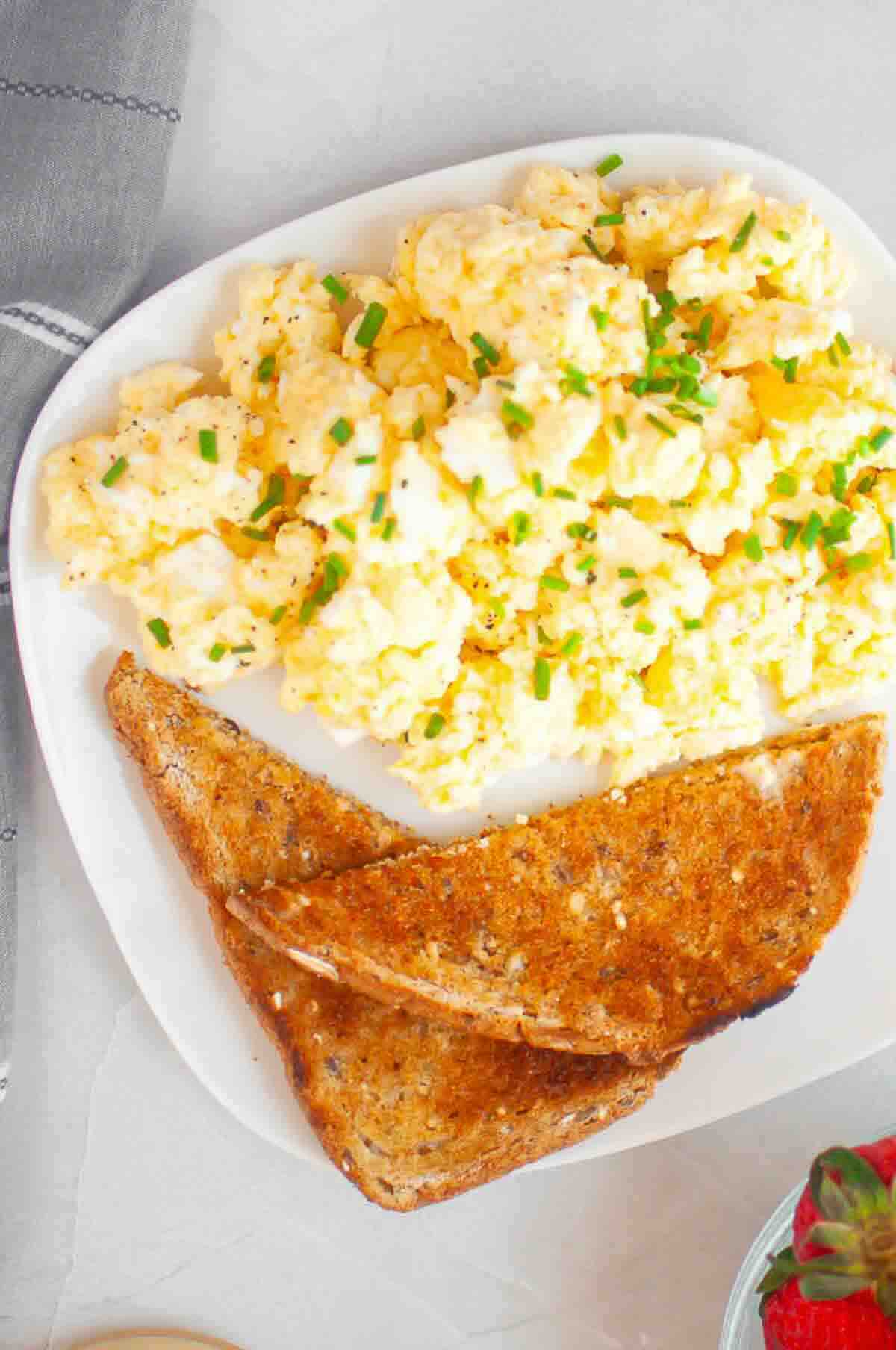 A white plate with scrambled eggs topped with chopped chives, served with two slices of toasted whole grain bread.