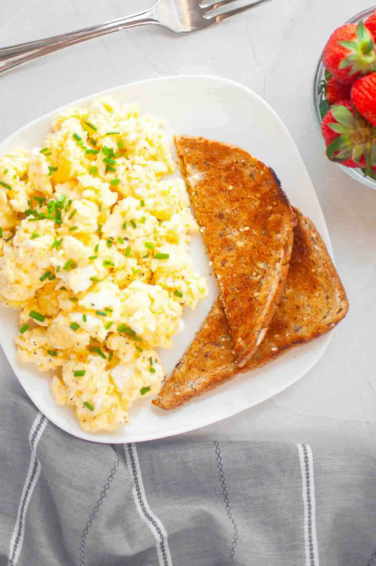 A plate with scrambled eggs garnished with chopped chives, two slices of toasted whole grain bread, and a bowl of strawberries beside it.