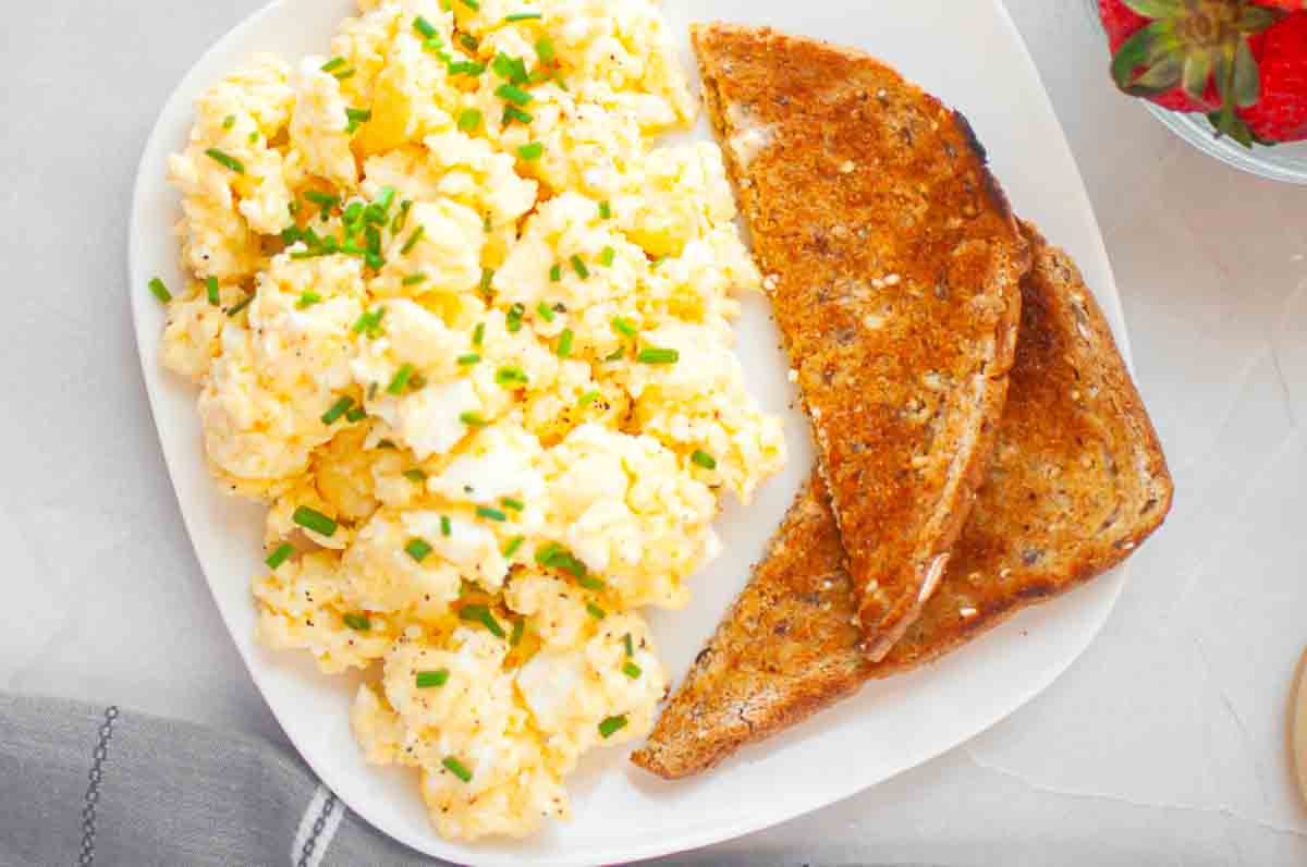 A plate with scrambled eggs garnished with chopped chives and two slices of toasted multigrain bread, with part of a bowl of strawberries visible in the corner.