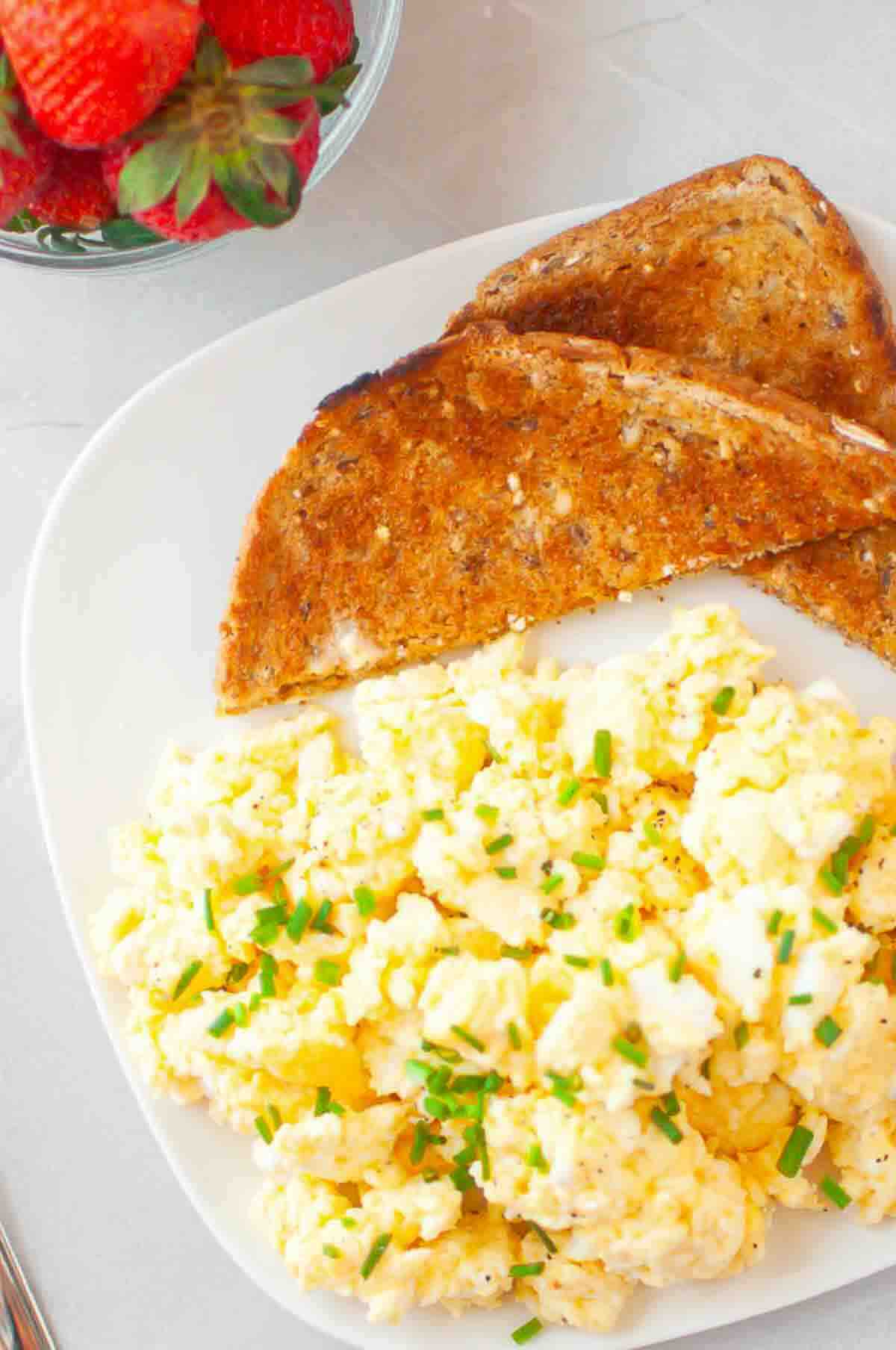 A plate with scrambled eggs topped with chopped chives and two slices of toasted bread, with a bowl of strawberries in the background.