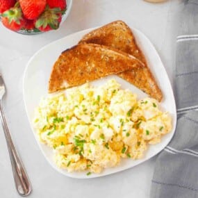 A plate with scrambled eggs garnished with herbs and two slices of toasted bread, next to a bowl of strawberries and a fork on a white surface.