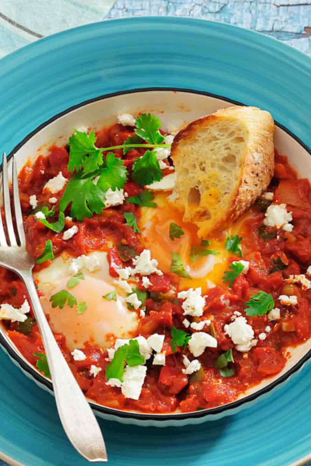 A bowl of shakshuka with poached eggs in tomato sauce, topped with feta cheese and cilantro, served with a slice of bread and a fork on a blue plate.