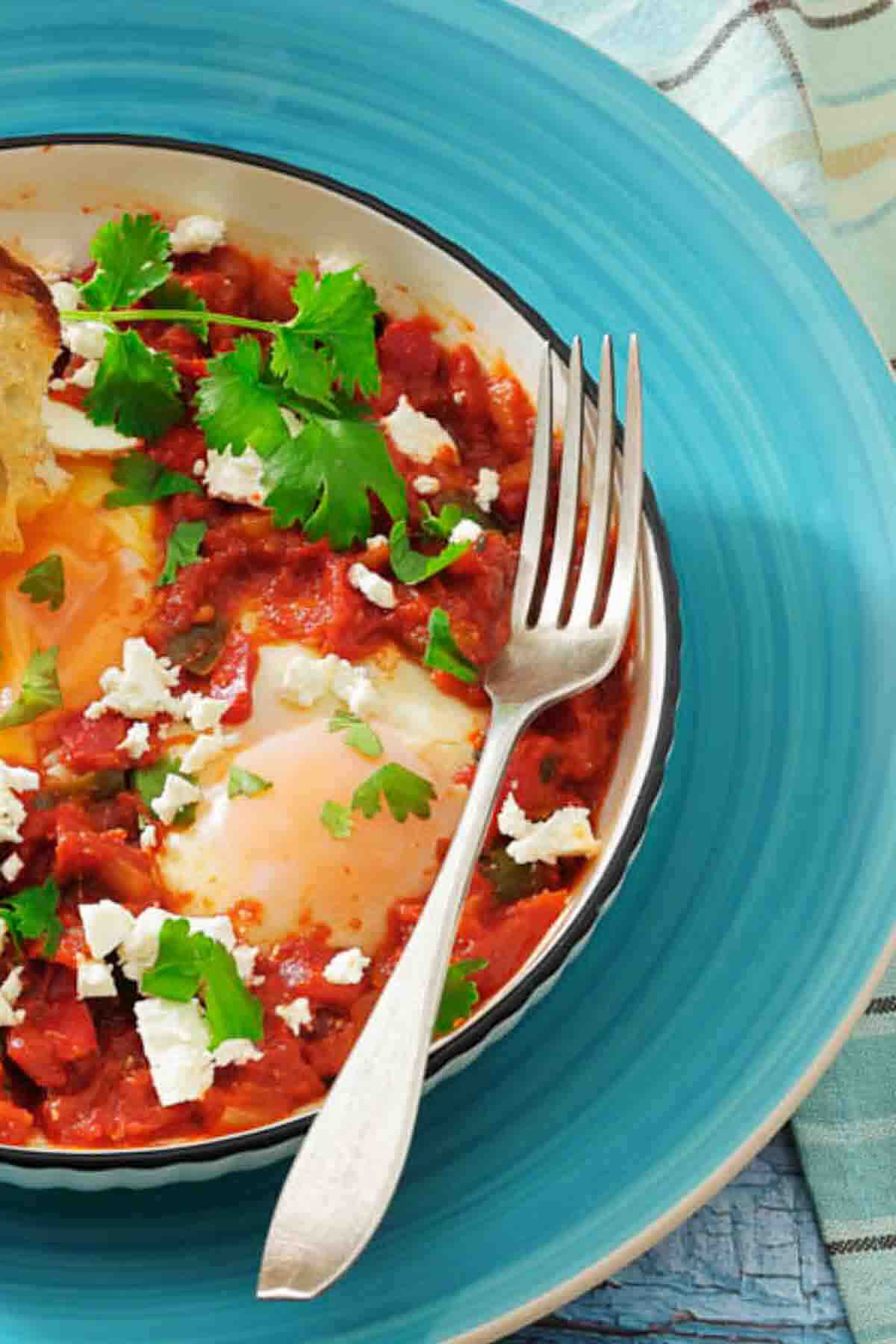 A bowl of shakshuka with poached eggs in tomato sauce, garnished with parsley and crumbled cheese, sits on a blue plate with a fork and a piece of bread.