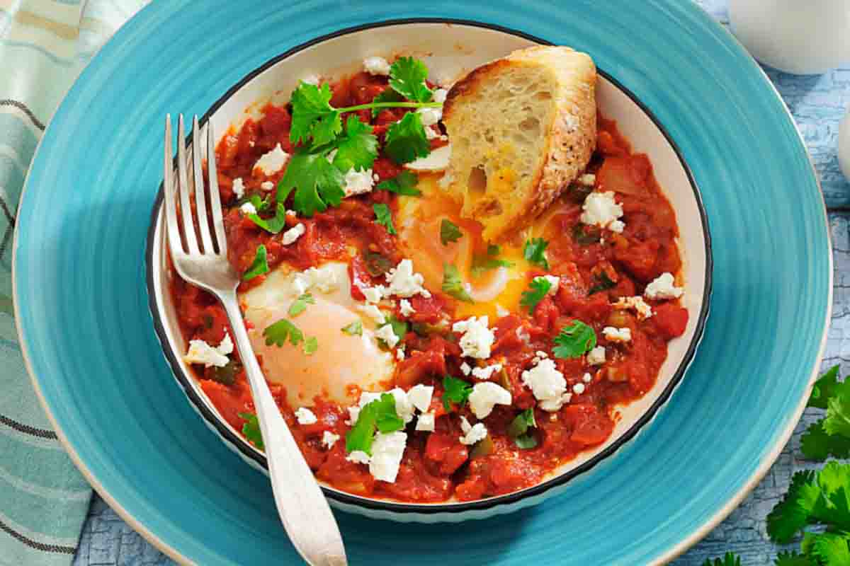 A bowl of shakshuka with poached eggs in tomato sauce, topped with herbs and cheese, served with a slice of bread and a fork on a blue plate.