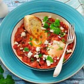 A bowl of shakshuka with poached eggs in tomato sauce, topped with crumbled cheese and herbs, served with toasted bread and a fork on a blue plate.