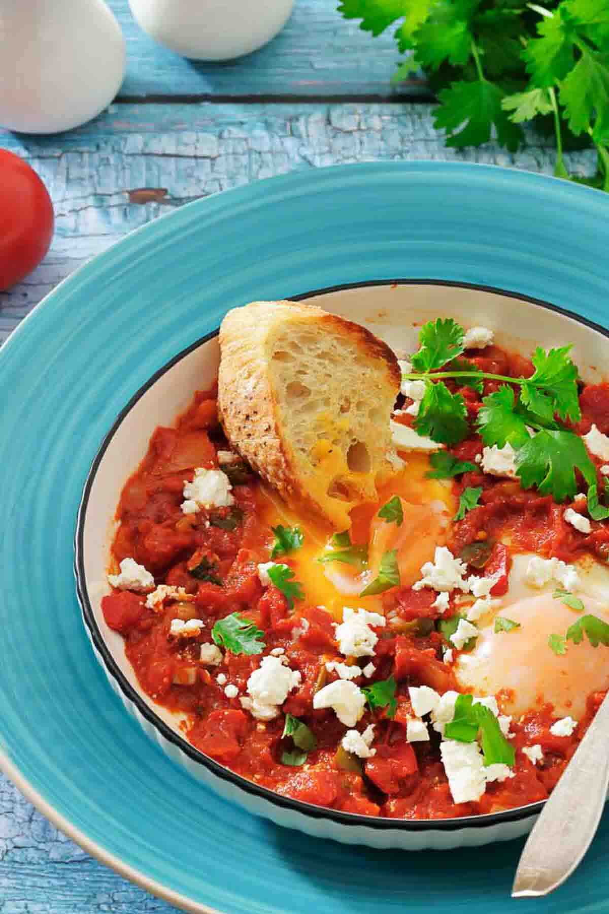 A bowl of shakshuka with poached eggs, tomato sauce, crumbled cheese, fresh herbs, and a slice of toasted bread on a blue plate.