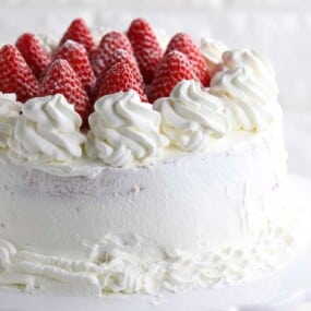 A round cake covered in whipped cream, topped with whole strawberries and decorative whipped cream swirls, displayed on a white cake stand.