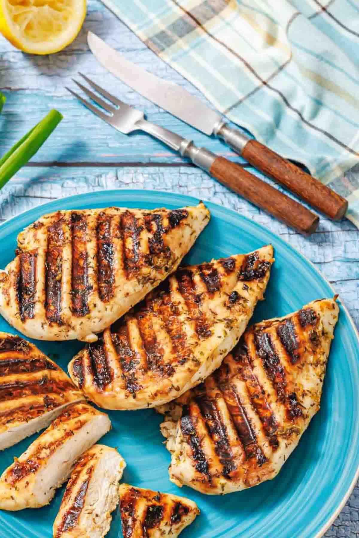 Grilled chicken breasts with visible grill marks are arranged on a blue plate, with utensils and a napkin in the background.