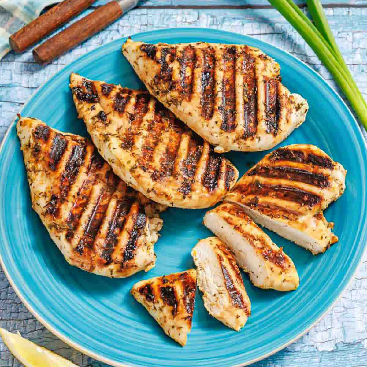 Grilled chicken breasts with char marks arranged on a blue plate, with one breast sliced, set on a light wooden surface.