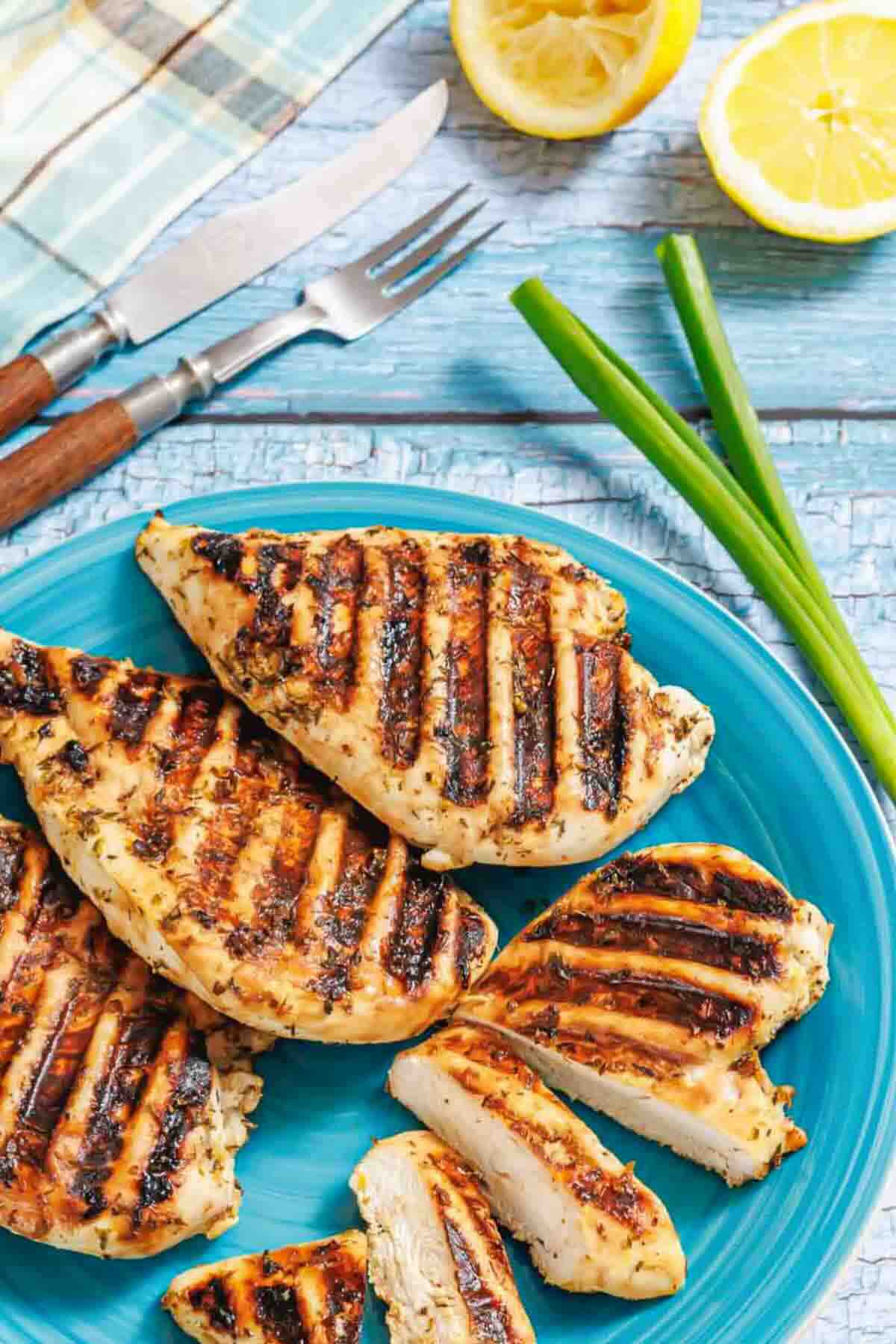 Grilled chicken breasts with grill marks on a blue plate, accompanied by green onions, lemon halves, and cutlery on a rustic blue wooden table.