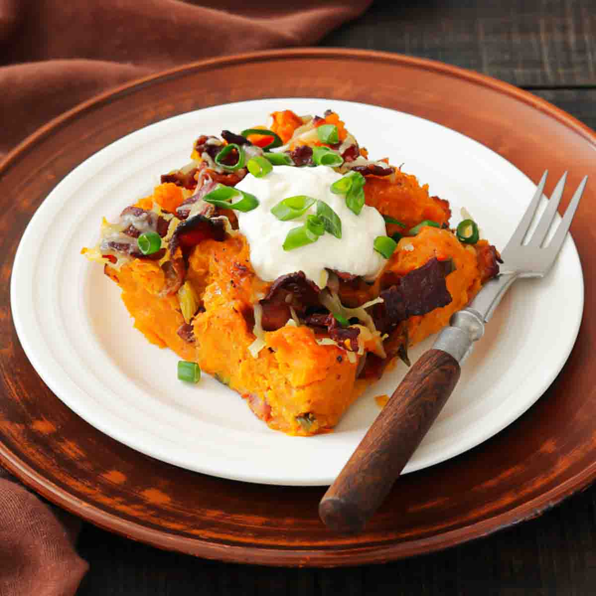 A serving of loaded sweet potato casserole topped with sour cream, chopped green onions, bacon, and cheese on a white plate with a fork.