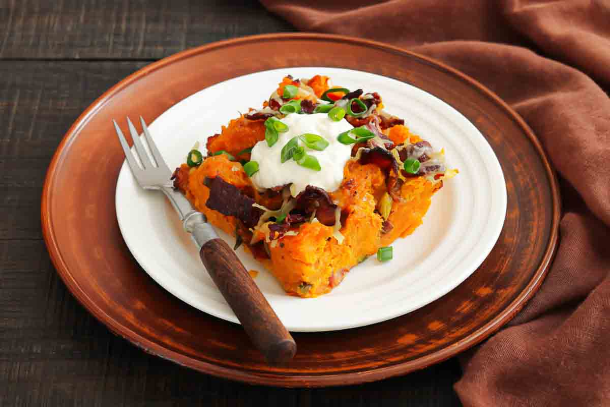 Square serving of sweet potato casserole topped with sour cream, chopped green onions, and bacon pieces on a white plate with a fork.