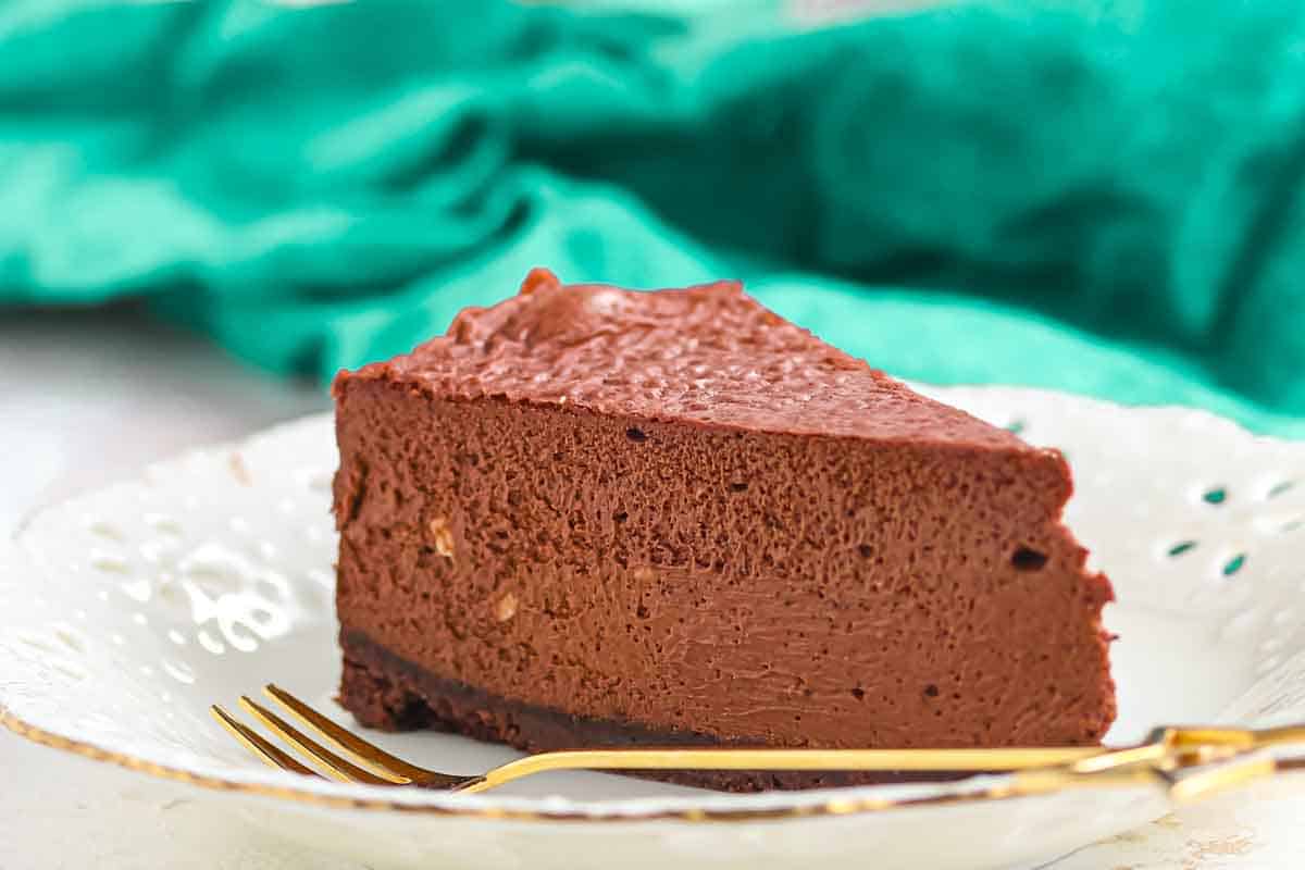 A slice of chocolate cheesecake on a white decorative plate with a gold fork, with a green cloth in the background.