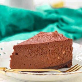 A slice of chocolate cheesecake on a white plate with a gold fork, set in front of a green cloth.