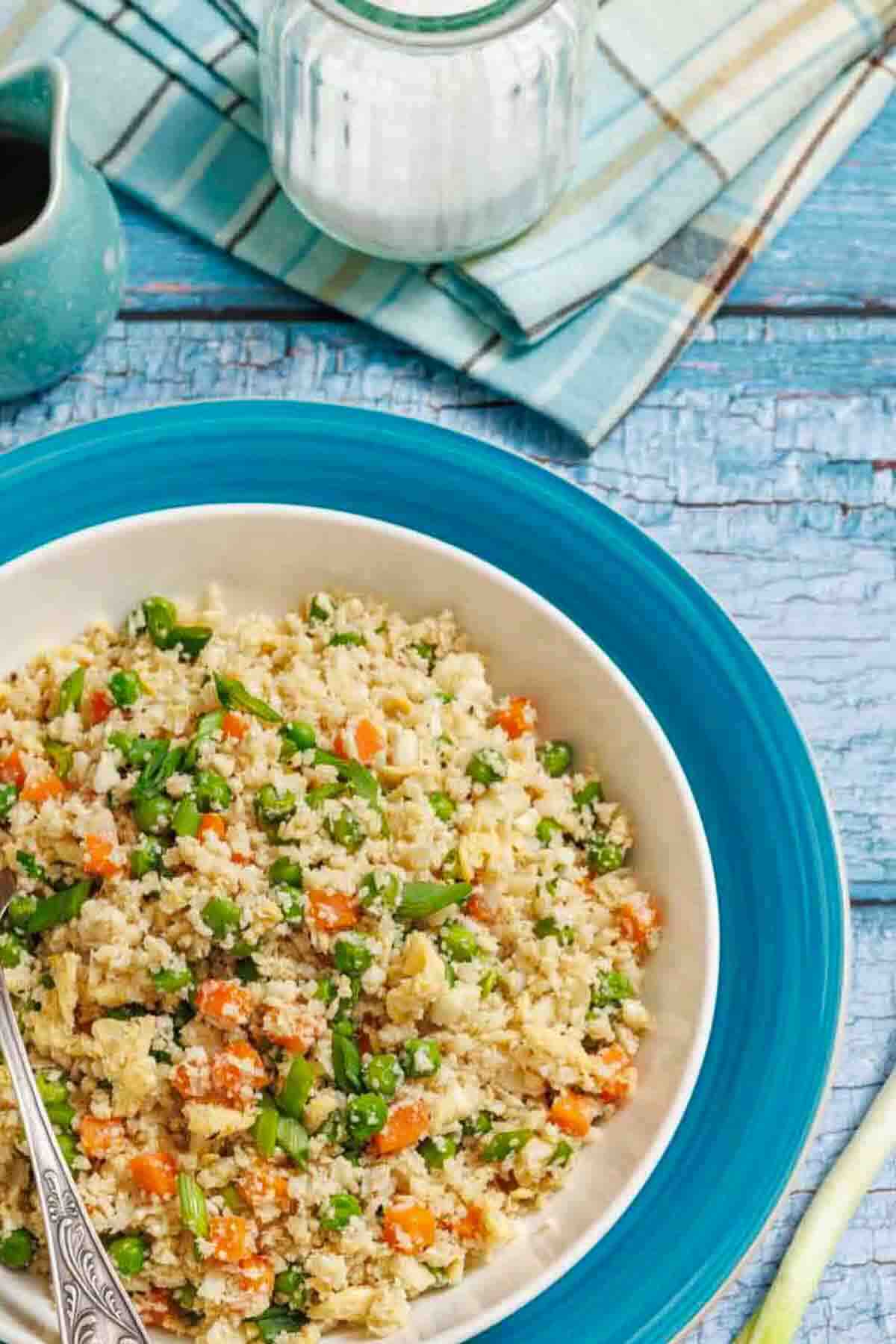 A bowl of cauliflower rice mixed with peas, carrots, and green beans sits on a blue plate, with a spoon in the bowl. A jar, napkin, and sauce are in the background.