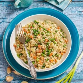 A bowl of vegetable couscous with a fork, placed on a blue plate atop a rustic blue wooden surface. Garlic cloves and green onions are nearby.