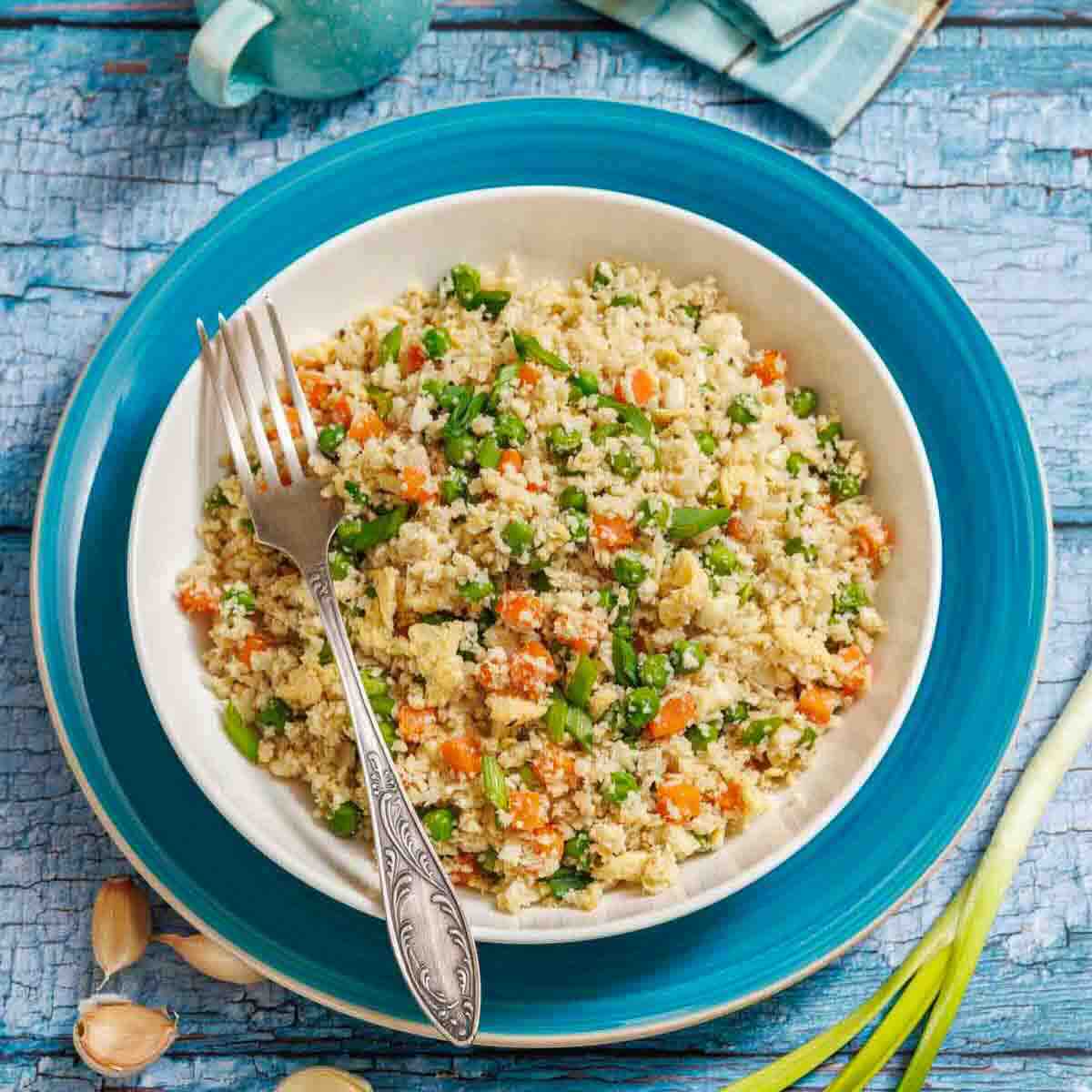 A bowl of vegetable couscous with a fork, placed on a blue plate atop a rustic blue wooden surface. Garlic cloves and green onions are nearby.