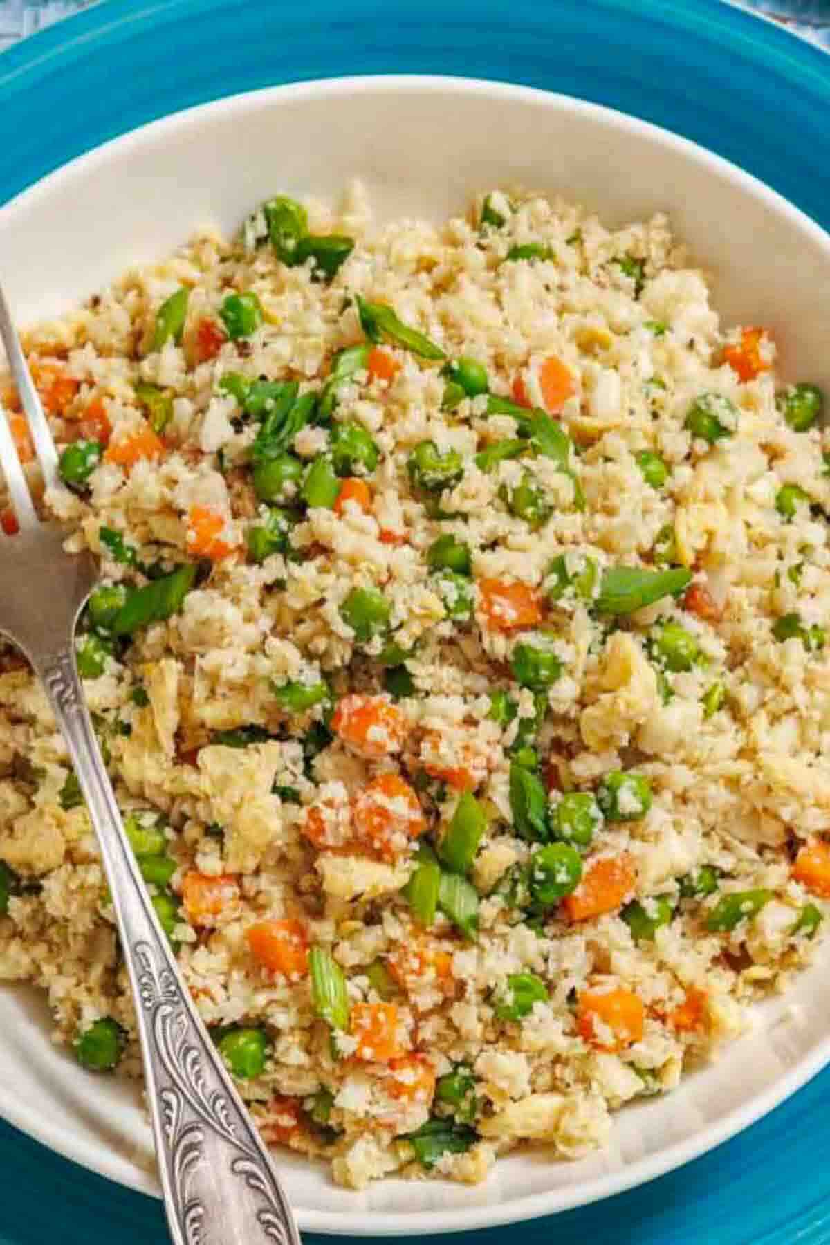 A bowl of cauliflower fried rice with peas, carrots, green onions, and scrambled eggs, served with a fork on a blue-rimmed plate.