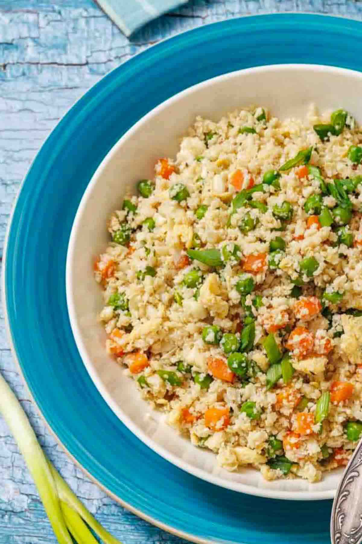 A bowl of cauliflower rice mixed with diced carrots, green peas, and chopped green onions on a blue plate, with a spoon on the side.