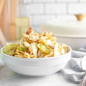 A white bowl filled with cooked cabbage, sliced sausage, and bacon pieces on a light-colored kitchen countertop.