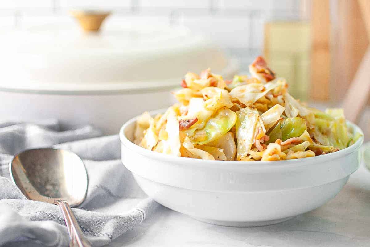 A white bowl filled with cooked cabbage, bacon, and sliced vegetables sits on a marble countertop next to a large spoon and a covered dish in the background.