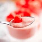 A close-up of a spoon holding pink mousse topped with small pieces of red fruit, with a blurred background.