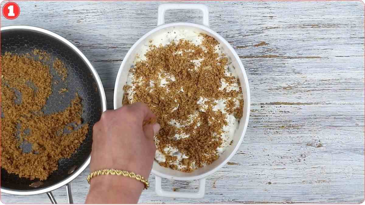 A hand sprinkles crumb mixture from a pan onto a white oval dish filled with a creamy layer, on a light wooden surface.