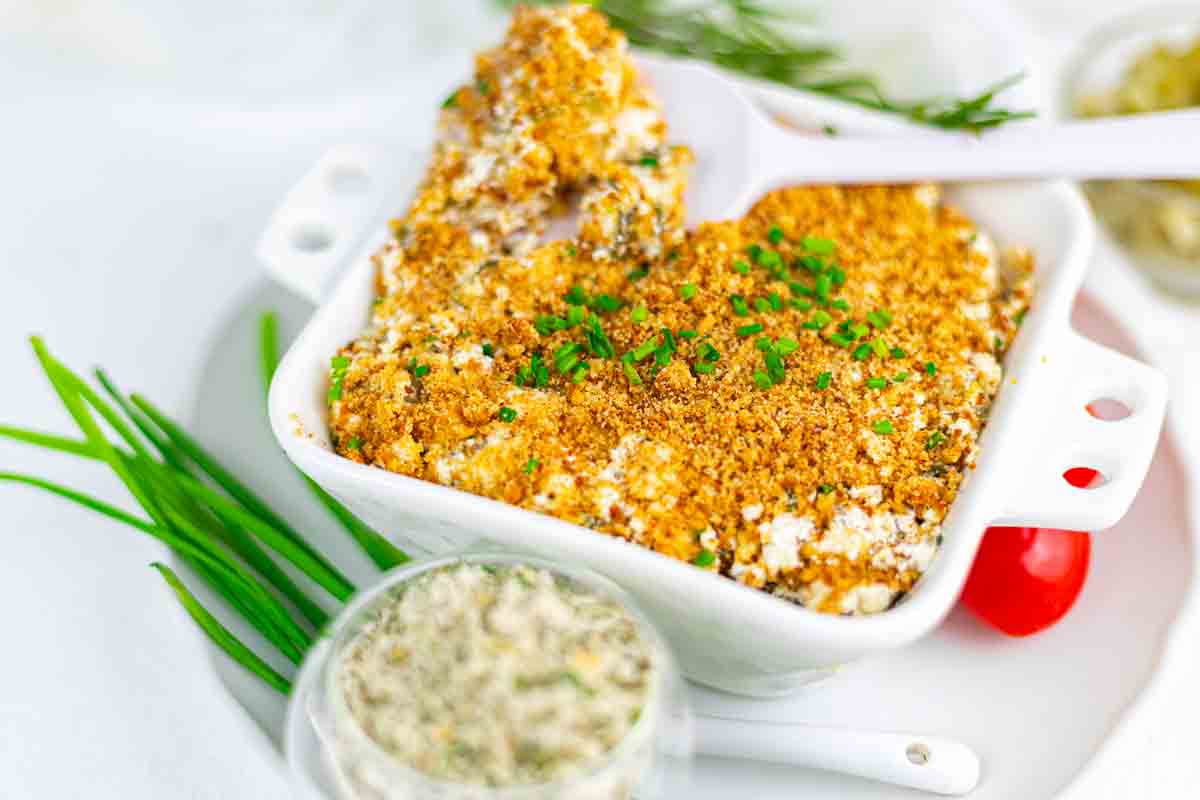 A baked casserole dip with a golden breadcrumb topping, garnished with chopped chives, served in a white square dish with a spoon.
