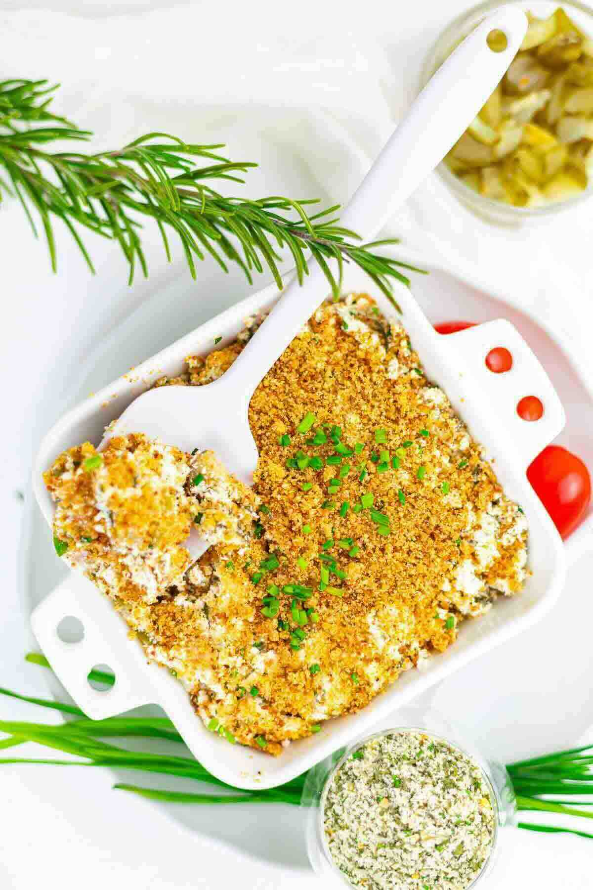 A white baking dish filled with a crumb-topped casserole, garnished with chopped herbs, with a white serving spoon and fresh herbs surrounding the dish.