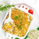 Baked casserole topped with golden breadcrumbs and chopped chives, served in a white dish with a white serving spoon. Fresh herbs and a seasoning mix are nearby.