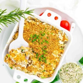 Baked casserole topped with golden breadcrumbs and chopped chives, served in a white dish with a white serving spoon. Fresh herbs and a seasoning mix are nearby.