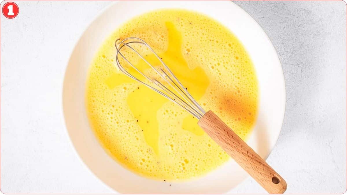 A metal whisk with a wooden handle rests in a bowl of beaten eggs on a light-colored surface.