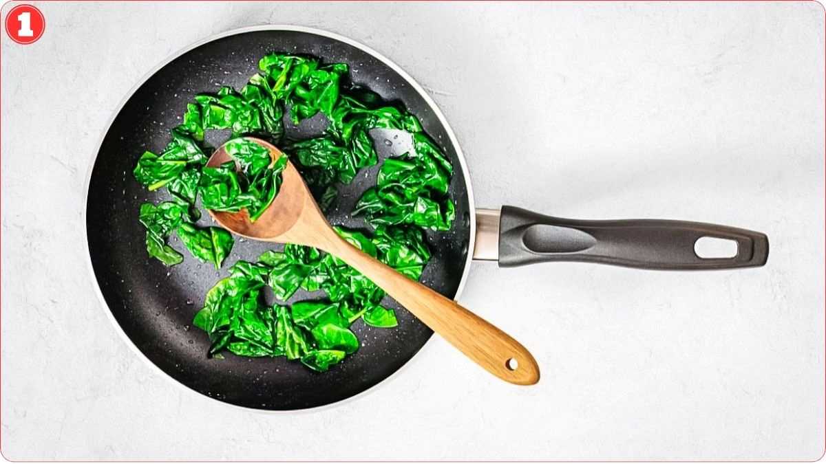 A nonstick frying pan with sautéed spinach and a wooden spoon on a light-colored surface.