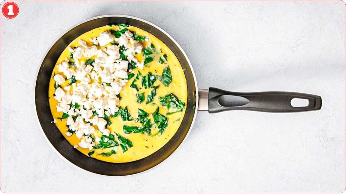 A frying pan contains an uncooked spinach and feta omelette mixture, viewed from above on a light surface.